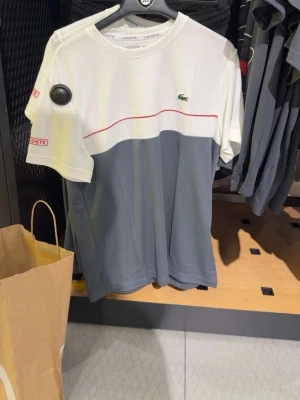 Vit och grå t-shirt från Lacoste - Snygg t-shirt från Lacoste i vitt och grått med en tunn röd linje över bröstet. Klassisk krokodil-logga på bröstet och rund hals. Perfekt för en clean och sportig look.