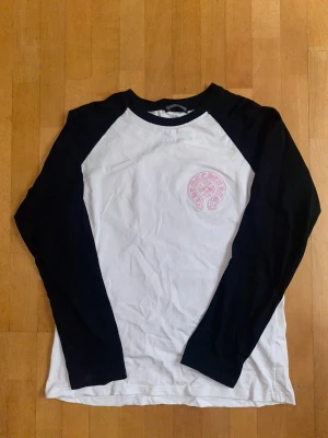 Svartvit långärmad tröja Chrome Hearts - Säljer en svartvit långärmad tröja från Chrome Hearts med svart krage och ärmar. Framsidan har ett rosa broderat Chrome Hearts-märke på bröstet. Tröjan har en klassisk baseballmodell och är gjord i mjuk bomull.