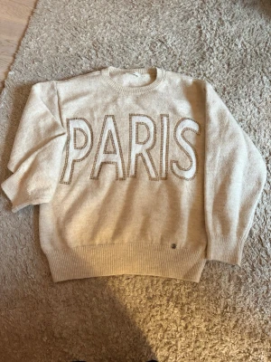 Beige stickad tröja med PARIS-tryck - Snygg beige stickad tröja med stor text 'PARIS' framtill i kontrasterande färg. Tröjan har rund halsringning, ribbade muddar och avslut. Perfekt för dig som gillar en avslappnad och trendig stil.
