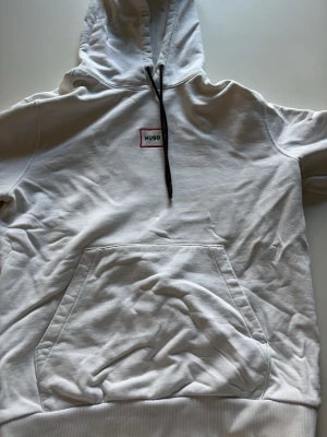 Vit hoodie från Hugo boss med magficka - Snygg vit hoodie från Hugo Boss med svart dragsko i huvan och en liten rödvit logga på bröstet. Hoodien har en stor magficka och ribbade muddar vid ärmsluten. Perfekt för en avslappnad och stilren look.