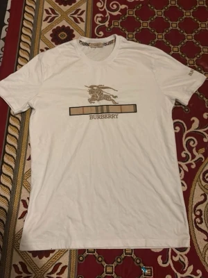 Vit Burberry t-shirt med logga - Säljer en vit t-shirt från Burberry med klassisk logga och riddartryck på bröstet samt Burberry-text på ärmen. T-shirten har rund hals och korta ärmar. Perfekt för dig som gillar stilrena och exklusiva plagg.