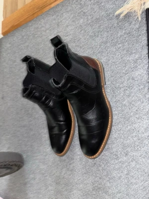 Svarta Chelsea boots med brunt parti - Säljer ett par stilrena svarta Chelsea boots i skinn med elastiska resårpaneler på sidorna och brunt parti vid hälen. Klassisk rundad tå och låg klack. Perfekta för dig som vill ha en tidlös och snygg look till hösten.