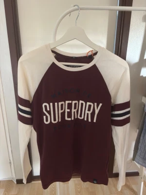 Vinröd och vit Vintage Superdry tröja - Snygg långärmad tröja från Superdry i vinrött med kontrasterande vita ärmar och detaljer. Tröjan har tryckt logga framtill och två vita ränder på ärmarna. Perfekt för en avslappnad och sportig stil.