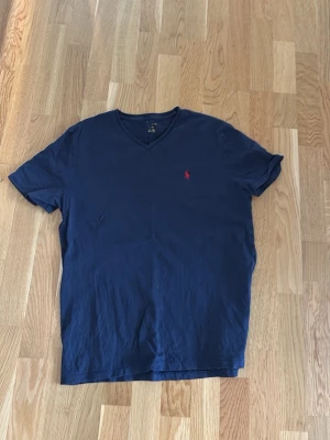 Marinblå v-ringad polo Ralph lauren - Snygg marinblå t-shirt med v-ringning och korta ärmar. Liten röd broderad logga på bröstet ger en clean look. Perfekt till jeans eller shorts för en enkel men stilren outfit.