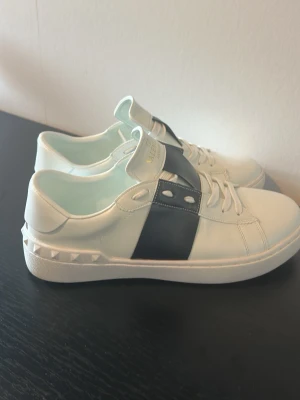 Valentino Garavani vita sneakers - Snygga vita sneakers från Valentino Garavani med bred svart detalj över mitten och nitar längs hälen. Klassisk låg modell med vita snören och guldfärgad logga på plösen. Skorna är i nyskick. Använda en gång. För små för mig. Köpte en storlek större. Säljer där av dessa. 