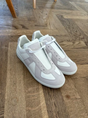 Mission Margiela white - Säljer ett par stilrena sneakers i vitt och beige med ovandel i slätt läder och detaljer i mocka. Klassisk låg modell med vita snören och gummisula. Perfekta för dig som gillar minimalistisk och clean stil.