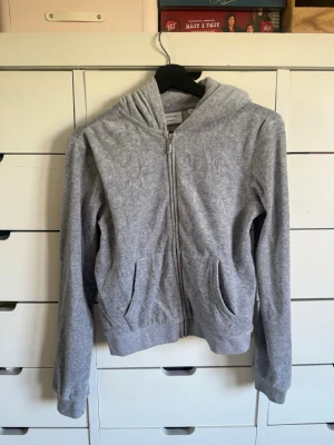 Grå zip hoodie med broderad logga - Säljer en grå hoodie med dragkedja och huva. Tröjan har två fickor framtill och en broderad logga på bröstet. Tillverkad i mjukt sweatshirtmaterial som är perfekt för chill dagar. Passar dig som gillar enkel och clean stil.
