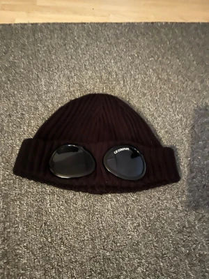 Svart goggle-mössa från C.P. Company - Säljer en svart ribbstickad mössa från C.P. Company med ikoniska svarta goggle-linser framtill. Mössan är tillverkad i mjukt material och har en uppvikt kant. Perfekt för dig som vill sticka ut med en streetwear-inspirerad look.