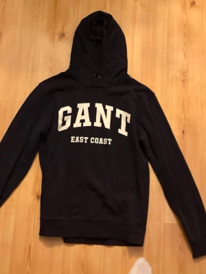 Marinblå hoodie från GANT med tryck - Snygg marinblå hoodie från GANT med stor vit text 'GANT EAST COAST' på bröstet. Klassisk modell med huva och dragsko. Perfekt för en avslappnad och trendig stil. Mjuk bomullskänsla och ribbade muddar vid ärmar och nederkant.
