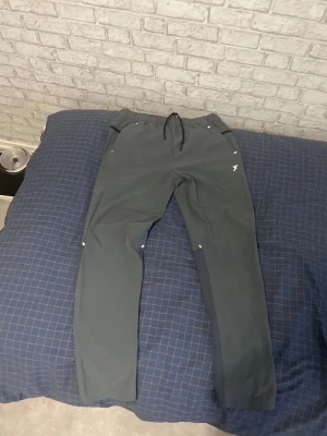 Grå byxor med dragkedjor+reflex - Snygga mörkgrå mjukisbyxor med elastisk midja och dragsko. Byxorna har raka ben och praktiska fickor på sidorna. En vit logga finns tryckt på ena benet. Perfekta för en avslappnad och sportig stil. OBS! STORLEK M MEN FINNS I S OCKSÅ! pris kan diskuteras.