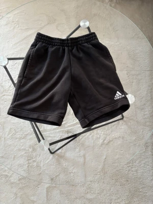 Svarta shorts från Adidas - Säljer ett par svarta shorts från Adidas med klassisk logga broderad i vitt på vänster ben. Shortsen har elastisk midja och två sidofickor. Perfekta för träning eller chill dagar. Tillverkade i mjuk bomull för skön känsla.