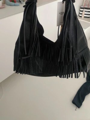 Svart fransväska från Ulrika - Snygg svart handväska från Ulrika med coola fransdetaljer och mjukt konstläder. Väskan har en rundad form och bred axelrem, perfekt för dig som gillar en trendig och bohemisk look. Enkel att matcha till olika outfits och rymmer det viktigaste.