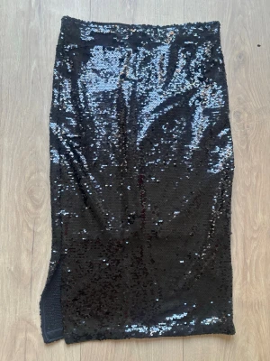 Svart paljettkjol med slits - Säljer en svart långkjol täckt med glittriga paljetter. Kjolen har en rak passform och en snygg slits på sidan som ger extra edge. Perfekt för dig som vill sticka ut och glänsa lite extra.