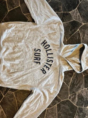 Grå hoodie från Hollister Surf - Snygg grå hoodie från Hollister Surf med boxy fit och stor magficka. Hoodien har broderad text i mörkblått framtill och en klassisk huva. Perfekt för chill dagar och streetwear-stil.