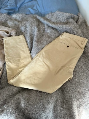 Beige chinos med slim - Snygga beige chinos med rak passform och klassisk design. Byxorna har en bakficka med knapp och är tillverkade i ett mjukt bomullstyg som känns skönt mot huden. Perfekta för en clean och stilren look.