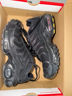 Nike Air Max Plus svarta sneakers - Säljer ett par svarta Nike Air Max Plus sneakers med mesh och syntetpaneler. Skorna har synliga Air-enheter i sulan och detaljer i gult på yttersulan. Klassisk TN-design med vågiga linjer och robust look. Perfekta för dig som gillar streetwear.