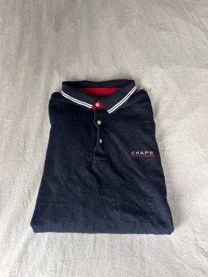 Mörkblå/svart långärmad pikétröja Chaps Ralph Lauren Ralph Lauren - Svart/mörkblå långärmad pikétröja från Chaps Ralph Lauren med vitrandig krage och röd insida. Klassisk pikéknäppning framtill och broderad logga på bröstet. Perfekt för en sportig och stilren look.