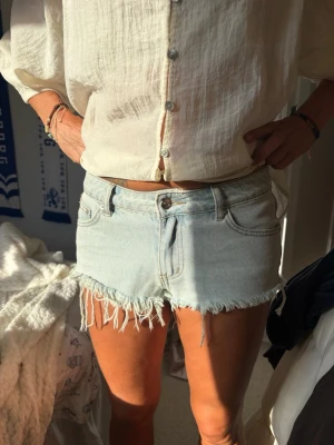 Ljusblå denimshorts med fransar - Säljer ett par ljusblå denimshorts med råa, fransiga kanter och klassisk femficksdesign. Shortsen har låg midja och är perfekta för varma sommardagar.                                                     Nypris =1100kr därav priset.                            SÄLJER HELT OANVÄNDA EFTERSOM DE E FÖR SMÅ