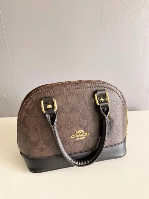 Brun och svart handväska från Coach - Snygg handväska från Coach i brunt och svart konstläder med guldfärgade detaljer. Väskan har Coach-logga framtill, två handtag och rundad form. Botten är förstärkt med metallfötter för extra hållbarhet. Perfekt accessoar för en trendig look.