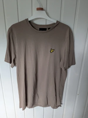 Beige t-shirt från Lyle & Scott - Snygg beige t-shirt från Lyle & Scott med klassisk gul logga på bröstet. T-shirten har rund halsringning och korta ärmar. Tillverkad i mjuk bomull för skön känsla och enkel stil.