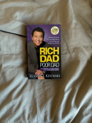 Rich Dad Poor Dad pocketbok - En engelskspråkig pocketbok med titeln 'Rich Dad Poor Dad' av Robert T. Kiyosaki. Omslaget är lila och svart med gul och vit text samt ett foto på författaren. Boken handlar om privatekonomi och vad rika lär sina barn om pengar.
