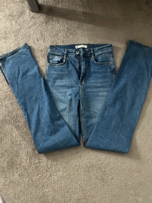 Blå raka jeans med hög midja - Säljer ett par klassiska blå jeans med raka ben och hög midja. Jeansen har slit nere, bälteshällor och stängs med knapp och dragkedja. Perfekta till alla dina outfits och superenkla att styla.