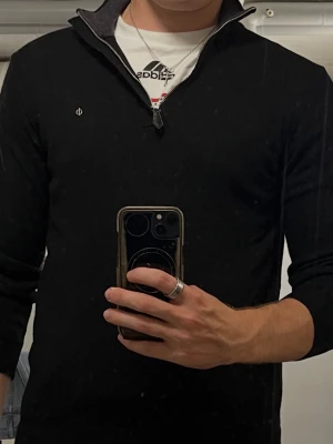 Svart stickad tröja med halv dragkedja - Säljer en svart stickad tröja med halv dragkedja och liten broderad logga på bröstet. Tröjan har ribbade muddar vid ärmslut och nederkant samt hög krage. Perfekt för lager på lager och enkel att matcha med olika outfits. Passar mera som S