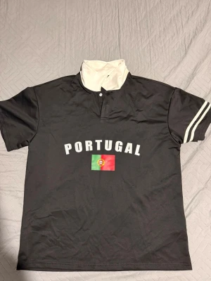 Svart Portugal t-shirt med tryck - Säljer en svart t-shirt med vit krage och tryckknappar. På bröstet står det 'PORTUGAL' i vitt och under finns Portugals flagga. Ärmarna har vita ränder som detalj. Perfekt för dig som vill visa ditt stöd för Portugal.