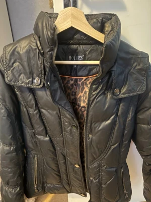 Svart pufferjacka från Hollies - Snygg svart pufferjacka från Hollies med hög krage, dragkedja och knappar framtill. Jackan har två fickor med dragkedja och coolt leopardmönstrat innerfoder. Perfekt för kalla dagar och ger en trendig look.Dragkedjan är sönder