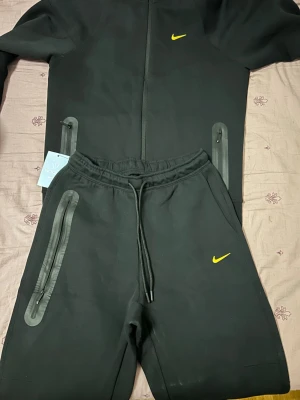 Svart Nike träningsset med huva - Storlek L. Säljer ett svart träningsset från Nike med gul logga. Setet består av en hoodie med dragkedja och huva samt matchande byxor med dragsko i midjan. Båda plaggen har dragkedjeförsedda fickor med svarta detaljer. Perfekt för träning eller chill.