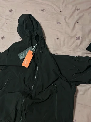Cp jacket Svart vindjacka med huva och dragkedja - Storlek L. Svart vindjacka med huva och hel dragkedja framtill. Jackan har långa ärmar, flera fickor med dragkedja och justerbar snörning i huvan. Perfekt för blåsiga dagar och enkel att matcha med olika outfits.