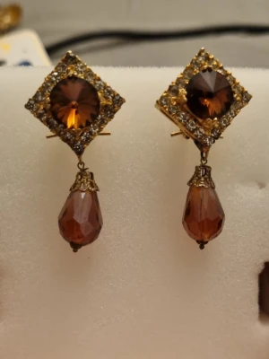 Bruna droppformade örhängen - Unika örhängen med fyrkantig guldfärgad ram, glittrande stenar och stora bruna droppformade pärlor. Örhängena har vintagekänsla och detaljerade metallinfattningar. Perfekta för dig som gillar statement-smycken och vill sticka ut.