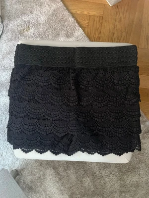 Svarta spetsiga shorts med bred midja - Säljer ett par svarta shorts med bred, elastisk midja och snyggt spetsmönster i lager på lager. Shortsen har en feminin look med vågig kant nertill och är perfekta för dig som gillar detaljer och stilren design. Ungefär storlek S/M