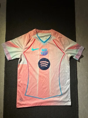 Barca fotbollströja - Säljer en snygg rosa Barca matchtröja från Nike med blå detaljer och klubbmärke på bröstet. Tröjan har V-ringning, korta ärmar och ett stort Spotify-tryck på magen. Perfekt för dig som älskar fotboll och vill sticka ut på planen.