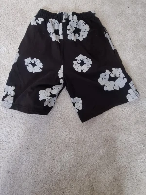 Svarta shorts med blommigt mönster - Säljer ett par svarta shorts med stora vita blommor. De har elastisk midja och avslappnad passform, perfekta för sommardagar. Materialet känns mjukt och lätt, troligen bomull eller polyester. Snygga och bekväma för en chill look.