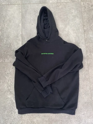 Svart hoodie Distorted Universe - Svart hoodie med huva och trycket 'DISTORTED UNIVERSE' i grönt på bröstet. På ryggen finns ett stort futuristiskt motiv med glitch-effekt och text. Hoodien har känguruficka och ribbade muddar. Perfekt för dig som gillar streetwear och unika prints. Storlek M 