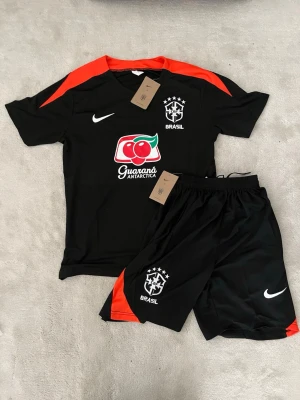  Brasilien träningset Nike - Snyggt svart träningsset från Nike med Brasilien-logga och Guaraná Antarctica-tryck. Tröjan har röda detaljer på axlarna och matchande shorts med samma logga och röd sidopanel. Perfekt för fotboll eller träning, tillverkad i lätt och snabbtorkande material.