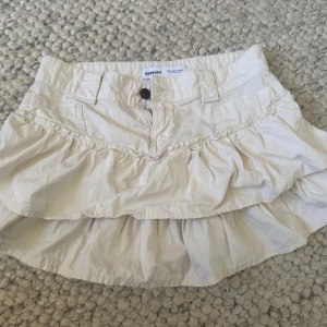 Beige jeanskjol  från Bershka - Söt beige kortkjol från Bershka med volangdetaljer och knapp framtill. 