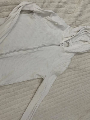 Vit ribbad hoodie med dragkedja H&M - Säljer en vit ribbad hoodie från H&M med dragkedja framtill och huva. Hoodien har långa ärmar och är tillverkad i ett mjukt, stretchigt material som är bekvämt att bära. Perfekt för en avslappnad och stilren look.