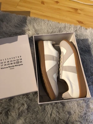 Maison Margiela vita sneakers med beige detaljer - Maison Margiela sneakers i vitt läder med beige mockadetaljer och beige snörning. Skorna har en klassisk gummisula i brunt och en stilren, retroinspirerad design. Perfekta för dig som gillar minimalistisk och trendig stil.