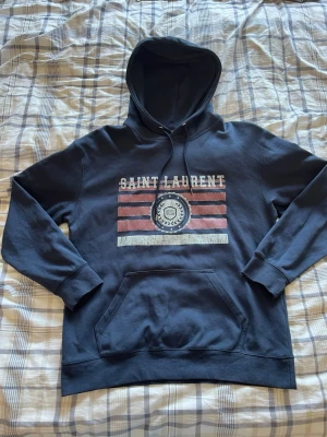 Mörkblå Saint Laurent hoodie - Säljer en snygg hoodie från Saint Laurent i marinblå färg. Den har ett vintageinspirerat tryck på bröstet med texten “Saint Laurent” och ett klassiskt “SL”-emblem. Hoodien har även luva med dragsko och en praktisk känguruficka framtill.  Detaljer:  * Märke: Saint Laurent * Storlek: M * Skick: Som ny * Färg: Marinblå med rött, vitt och blått tryck  En stilren hoodie i hög kvalitet som passar till det mesta.  Skickas snabbt och spårbart📦 