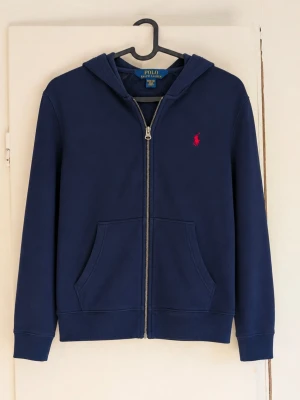 Ralph Lauren zip hoodie - Säljer en mörkblå zip hoodie från Polo Ralph Lauren med den klassiska röda loggan på bröstet. Hoodien är i väldigt fint skick och passar perfekt till både vardag och en mer uppklädd stil. Den är självklart äkta✅ Den är i bra skick och har inga fläckar eller skador😁