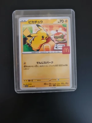 Pokémonkort Pikachu McDonald's promo - Unikt Pokémonkort med Pikachu från McDonald's samarbete, innesluten i plastficka. Kortet har japansk text och motiv av Pikachu vid ett bord med hamburgare och dryck. Perfekt för samlare av Pokémonkort och animeprylar.
