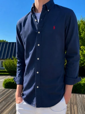 Mörkblå skjorta med knappar - Klassisk mörkblå skjorta med knappar framtill och button-down krage. Skjortan har lång ärm och en liten röd broderad logga på bröstet. Tillverkad i bomull och passar perfekt till jeans eller chinos.