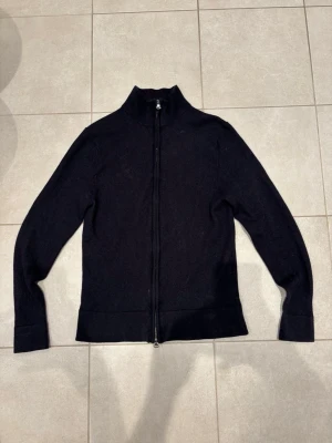 Mörkblå merinoull fullzip från These Glory Days - Mörkblå fullzip från These Glory Days i Superior Merino-ull. Tröjan har hel dragkedja framtill, hög krage och långärmad design. Stilren och enkel look som funkar till många outfits. Materialet är mjukt och värmande merinoull. S men sitter som XS. Ny pris 1000.
