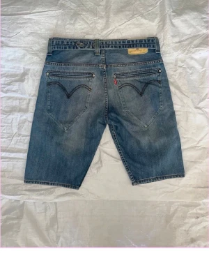 Blå jeansshorts med fem fickor - Klassiska blå jeansshorts i normal passform med fem fickor och knappgylf. Shortsens bakfickor har dekorativa sömmar och en liten röd etikett på höger sida. Perfekta för varma dagar och enkel att matcha med olika stilar.