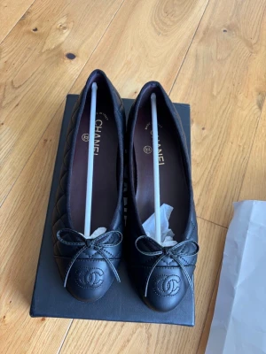 Svarta ballerinaskor från Chanel - Säljer ett par klassiska svarta ballerinaskor från Chanel i quiltat läder med rund tå och dekorativ rosett framtill. Skorna har den ikoniska Chanel-loggan på tån och elegant sömnad längs sidorna. Perfekta för dig som gillar tidlös och lyxig stil.
