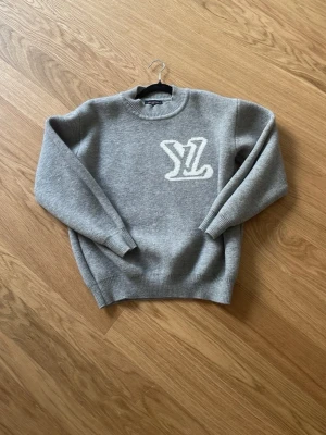 Grå stickad tröja med vit tryck - Mysig grå stickad tröja med rund hals och ribbade muddar. Stor vit 'YL'-logga på bröstet ger en cool och trendig look. Perfekt för dig som gillar enkel men snygg stil. Passar till jeans eller kjol för en avslappnad vibe.