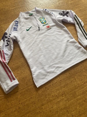 Vit Brasilien träningsoverall från Nike - Helt ny! Säljer en vit långärmad träningströja från Nike med Brasilien CBF-logga på bröstet. Tröjan har halv dragkedja, grönt Nike-märke, och tryckta sponsorloggor på ärmar och bröst. Perfekt för fotbollsträning eller som supporterplagg.