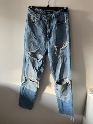 Blå ripped jeans från Missguided - Säljer ett par ljusblå jeans från Missguided med hög midja och stora slitningar på både lår och knän. Jeansen har raka ben och råa kanter vid bensluten. 